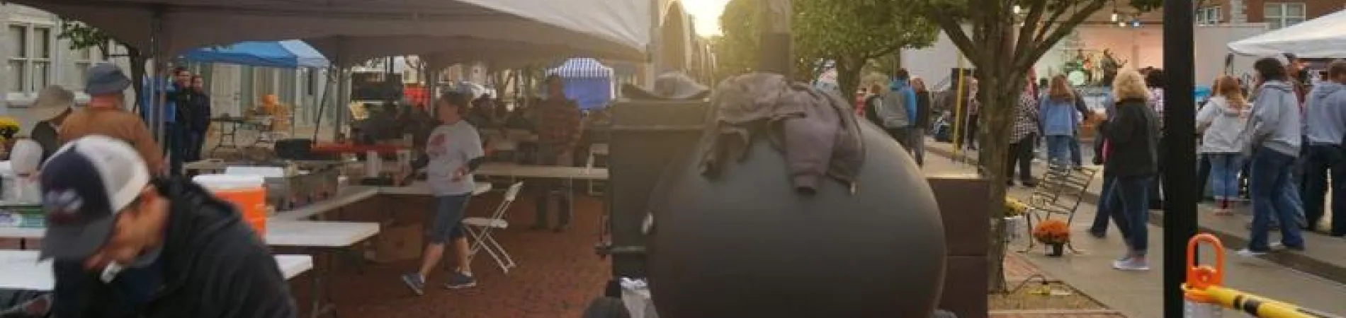 Street festival with tents, people, and a large BBQ grill at sunset.