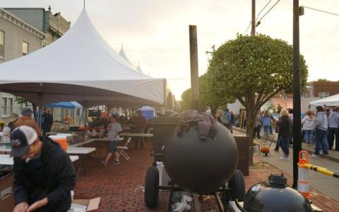 Street festival with tents, people, and a large BBQ grill at sunset.